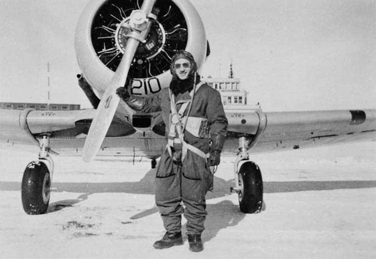 Photographie en noir et blanc – Un homme portant un lourd équipement de pilote se tient devant un avion, un bras déposé sur l’hélice.